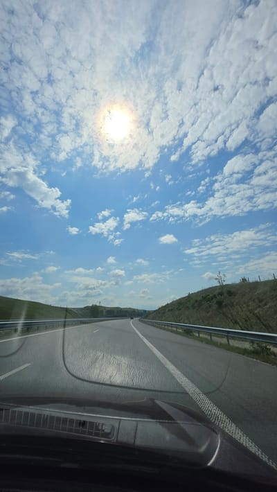 The image presents a view from inside a vehicle, possibly on a highway in Leshnitsa, Bulgaria. The road stretches out to the right, marked with white lines, next to a metal guardrail and grassy terrain. The sky occupies the left side of the frame, with scattered white clouds and a bright sun. Part of the vehicle's dashboard is visible in the lower right corner. There are no people immediately visible in the frame.