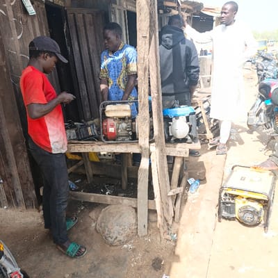 The image depicts an outdoor scene in Sabon Gari East, Nigeria, characteristic of a street-side repair or sales area. Four adult males are visible under daylight conditions.

In the left foreground, a male wearing a black baseball cap, a red short-sleeved t-shirt with a white stain on the front, dark trousers, and green "GUCCI" slides, stands leaning slightly forward, appearing to examine a small, dark object held in both hands. Directly behind him, positioned behind a rustic wooden workbench, a male in a blue and yellow patterned short-sleeved shirt stands, observing the first individual.

On the wooden workbench, two portable generators are present: a red and beige unit on the left and a blue and white unit on the right. Directly beneath the workbench, a large, dark, irregularly shaped stone object rests on the dusty ground, surrounded by scattered debris, including a discarded plastic bottle.

Further to the right, a male in a black hooded sweatshirt stands with his back partially towards the viewer, also positioned behind the workbench. To his immediate right, a fourth male, dressed in a white long-sleeved tunic and light-colored sandals, stands facing right, with his right arm raised and hand resting on an unseen surface or gesturing.

In the right foreground, a yellow and black portable generator with a metal frame rests on the dirt ground. The background reveals additional street or market activity, including several motorcycles and rudimentary structures made of corrugated metal and wood. The ground throughout the scene is visibly dusty.