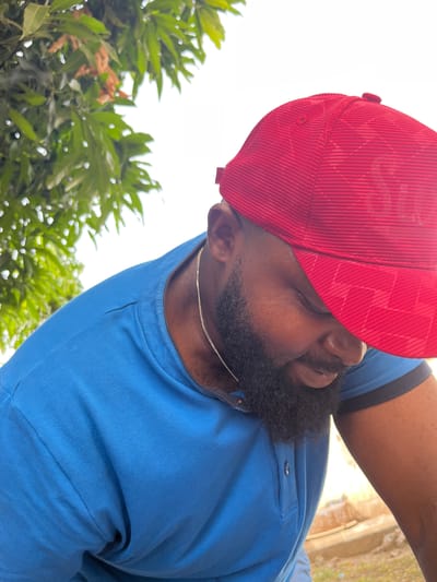 An outdoor, rotated close-up shot features a person, likely a man, facing away from the camera's direct line of sight. He has dark skin, a prominent black beard, and is wearing a bright red corduroy baseball cap, which appears to have a subtle plaid pattern on the crown. A silver chain necklace is visible around his neck. He is dressed in a short-sleeved blue polo shirt with a buttoned placket.
The image is brightly lit, indicating daylight, and the background is largely an overexposed white sky, suggesting a clear day. Below the person's head and to the left (due to the rotation), lush green foliage from trees or shrubs is visible, some of which are slightly out of focus, adding depth to the scene. The overall impression is a casual, relaxed moment captured outdoors. No text is visible in the image.