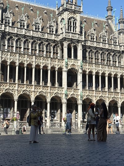 This image captures a daytime scene in the Grand Place, Brussels, Belgium, showcasing the iconic Gothic architecture of the Town Hall. In the foreground, a cobblestone square is visible with several individuals. A woman wearing a headscarf and a yellow top stands in the center, holding a camera and taking a photo. To her right, two women are standing and looking at a phone. Further back, on the left side of the image, a man in a white t-shirt and shorts walks away from the camera. On the right side, another man in a white t-shirt and shorts stands, facing towards the building. The background is dominated by the ornate facade of the Town Hall, characterized by its numerous arches, columns, statues, and intricate stonework. The sky is clear and blue, indicating fair weather.