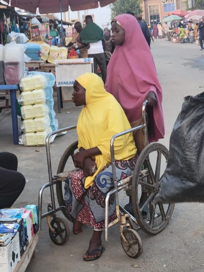 Here's a detailed description of the image:

**Setting:** An outdoor market scene in Madalla, Nigeria.

**Subjects:** Two African women are the primary focus, one wearing a yellow headscarf and the other a pink one. Both are sitting on an old, metal-framed cart with wheels. A third person, wearing black pants, is partially visible in the foreground. There are several other people visible in the background, further back in the market.

**Objects:** The cart is loaded with various items including rectangular packages wrapped in clear plastic. Additional merchandise, including packaged food items, is arranged on a shelf next to the cart. The street and buildings form the background.

**Actions and Interactions:** The woman in the yellow headscarf is sitting on the cart and appears to be holding her knees. The woman in the pink headscarf is behind her. The cart appears to be part of their market setup. The other people in the background are likely shoppers or vendors.

**Environment:** The location is a busy marketplace with various vendors and shoppers. The ground appears to be paved. The lighting suggests it is daytime.