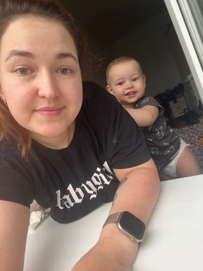 A woman and a toddler are in an indoor setting, possibly a bedroom, with a view of a window. The woman is in the foreground, taking a selfie. She has brown hair and is wearing a black t-shirt with white lettering. Her left arm, with a smartwatch, is visible. The toddler, positioned behind her, is facing the camera and smiling. The toddler has fair hair and is wearing a grey t-shirt with cartoon graphics and a diaper. The background includes a bed with patterned bedding and a dresser with drawers. The environment appears to be a domestic space. The provided image does not contain any information that would indicate the location is Chelyabinsk, Russia.