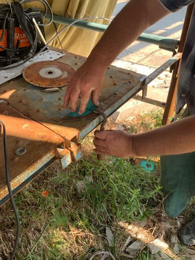 A close-up, overhead shot captures two gloved hands working on a rusty, turquoise metal workbench outdoors. The workbench is cluttered with various items, including a circular saw blade with a rusted edge, a welding machine, and a blue-green cutting disc. One hand is holding a tool with a curved metal end, seemingly manipulating the cutting disc. The other hand is positioned over the workbench, its fingers close to the disc. The scene is set against a backdrop of dry grass, some green foliage, and fragments of what appear to be construction debris. A small red flower is visible amongst the grass. The lighting suggests it is daytime, with dappled sunlight creating shadows. This scene is located in Pivdennoukrainsk, Ukraine, with the environment indicating an outdoor workshop or construction area.