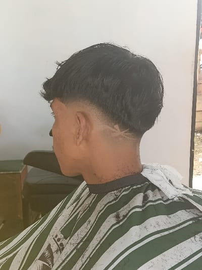 A male individual is depicted in right profile, seated and receiving a haircut in Mns Alue, Indonesia. He has short, dark hair, with the sides and back faded and a stylized geometric design shaved into the hair on the right side of his nape. The subject is wearing a green and white vertically striped barber's cape, secured at the neck with a white disposable paper collar. Numerous small, black hair clippings are visible on the cape and scattered around the subject's neck and ear. In the background, a dark, upholstered barber's chair is partially visible to the left of the subject, positioned against a plain white wall.