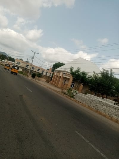 The image depicts a street scene in Kuson, Nigeria. A paved road, marked with a white line, dominates the foreground and slopes upwards and to the right. On the left side of the road, two yellow auto-rickshaws, known locally as Keke NAPEPs, are driving away from the camera. The Keke NAPEP in front has the number "7147" visible on its rear.

Further down the road on the left, a modern building with multiple windows and light-colored walls is visible. To the right of the road, a distinctive building with a large, white, pyramidal roof stands prominently. This building has a light brown facade with columns and windows. In front of this building, a low wall constructed from light-colored bricks runs along the roadside. Trees with dense green foliage are planted between the road and the building, and also further to the right. The background shows a hilly landscape under a partly cloudy sky.

The lighting suggests it is daytime, likely late morning or afternoon, given the bright but diffused light and the presence of clouds. The weather appears to be fair. The scene is characterized by urban infrastructure and typical modes of transportation in Nigeria. There are no people clearly visible in the image, but the presence of the vehicles indicates ongoing activity. The overall impression is one of a normal day in a Nigerian town.