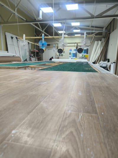 The image depicts an indoor workshop or studio space. In the foreground, a wooden surface, possibly a workbench or table, is visible from a low-angle perspective. It appears to be a light-colored wood with a grain pattern and is dusted with white powder.

In the mid-ground, a long, raised structure covered with a green mesh material stretches across the width of the frame. This structure is supported by wooden framing. Several fans are suspended from the ceiling by chains, with one blue fan prominently positioned. Some fans have heat lamps attached to them. Behind this setup, a person is visible in the distance, appearing to be working or standing in the space. Various sheets of material, possibly fabric or paper, are leaning against a wall on the right side of the image. There are also shelves with items on them, and some framed artwork or prints are displayed on a wall in the background. The ceiling has exposed beams and ductwork, with bright fluorescent lighting panels illuminating the space. The overall impression is of an active workspace, possibly involved in a craft, manufacturing, or artistic process. There is no visible text in the image.