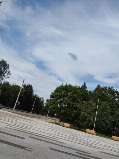 A tilted, full shot captures an outdoor scene with a paved area in the foreground and a line of trees and a road in the mid-ground, under a partly cloudy blue sky. The paved area is made of large, light-grey rectangular tiles, with darker grey rectangular patterns forming lines that recede into the distance. Several wooden benches with backrests are placed along the edge of the paved area, near the trees. One bench has a single person sitting on it, looking down. In the mid-ground, a road runs horizontally, bordered by a metal fence and grass. Beyond the road, a dense line of green trees forms a backdrop, with some taller trees visible in the distance. A lamppost stands on the left side of the paved area, near the road. The sky is a pale blue with wispy white clouds and a darker, small cloud formation towards the top center. The image has a slightly grainy texture. This scene is located in Kingisepp, Russia.