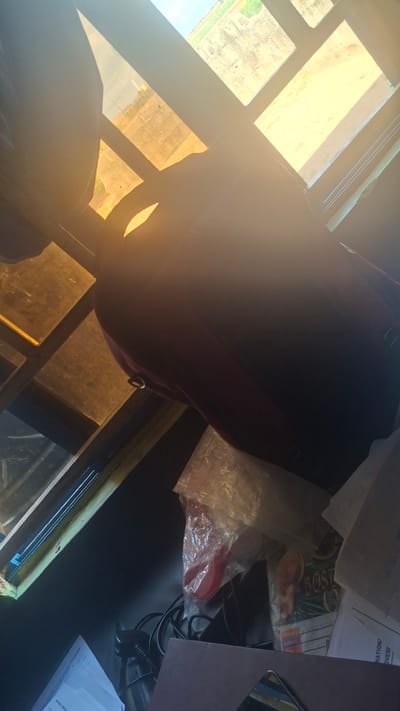 A dark red backpack rests on a table near a window with sunlight streaming through the panes. The window is divided into several sections, some of which show an outdoor scene with greenery and a faint hint of a road or path. The sunlight creates a bright glare in the image, particularly on the left side.

On the table, beyond the backpack, are various objects including papers, a clear plastic bag containing a red object and possibly other items, and some cables. One of the papers has visible text, including the word "NATION". Another object appears to be a magazine or book with the word "BOSTON" visible on it.

The scene appears to be indoors, likely an office or study area, given the presence of papers and the desk-like surface. The time of day is suggested by the strong sunlight, possibly late morning or early afternoon. The overall mood is neutral, with the focus on the objects and the interplay of light and shadow. The location is identified as Sunkani, Nigeria.