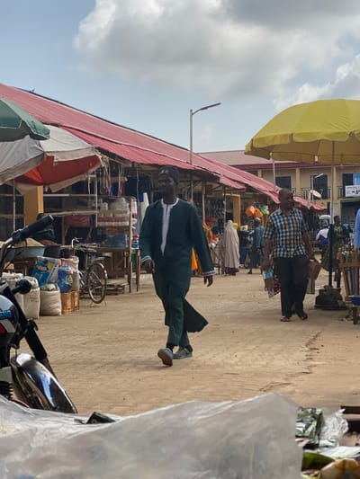 A man in a dark green robe and cap walks in front of shops under a red-roofed structure, lined with various items for sale, at what appears to be an open-air market in Kafanchan B, Nigeria. Another man in a plaid shirt walks in the background. There is a motorcycle parked in the foreground, partially covered by a white plastic sheet. Other items for sale include bags of goods and assorted household items. A yellow umbrella provides shade to the market area. The sky above is partly cloudy.
