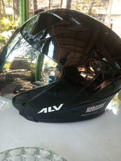 A black motorcycle helmet with a reflective visor is placed on a white surface. The helmet has a silver visor with a chrome accent. The word "ALV" is printed in white on the side of the helmet. Below that, a logo with the text "GARASIOT MOTOSHOP" and "DESIGN BY ALV HELMET" is visible. The background shows a slightly blurred view of foliage and what appears to be part of a building with a tiled roof, suggesting an outdoor setting in a residential or commercial area. This could be a scene captured in Tangerang, Indonesia, where such helmets are common for motorcycle transportation.