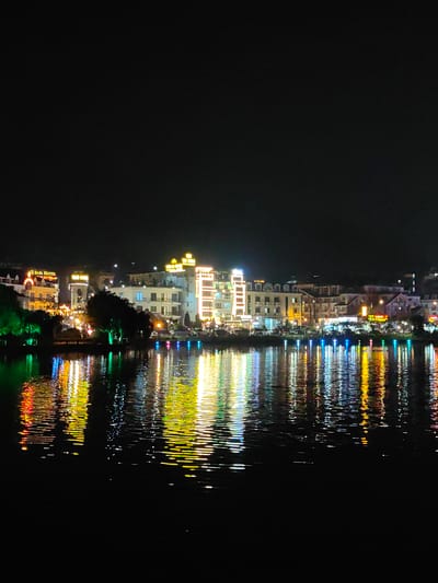 The image presents a nighttime wide view of the illuminated city shoreline of Sa Pa, Vietnam, seen across Sa Pa Lake. The upper third of the frame is an undifferentiated dark sky. Below, a continuous line of multi-story urban structures occupies the mid-ground along the lake's edge. These buildings feature numerous windows and are extensively lit by artificial sources, predominantly yellow, white, and orange light. Visible illuminated signage includes "O SAPA HOTEL" on the left and "PARK HOTEL" further left. Interspersed along the shoreline are trees and shrubs; some trees on the far left are illuminated with green light. At the immediate water's edge, a series of smaller, distinct multi-colored lights (blue, green, red, yellow) are visible. The lower two-thirds of the image comprises the dark, reflective surface of Sa Pa Lake, upon which the urban lights are cast as vibrant, elongated, and shimmering streaks, distorted by surface ripples. No human figures or discernible activities are present. The scene depicts an urban lakeside environment under artificial night illumination.