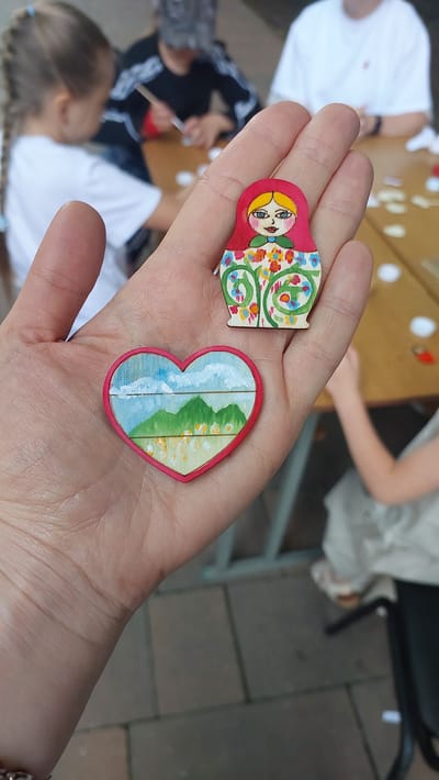 A person's open palm holds two small, hand-painted wooden ornaments. The ornament in the upper part of the palm is a stylized matryoshka doll with a red head, blue eyes, and a dress decorated with colorful flowers and green vines. Below the matryoshka is a heart-shaped ornament with a red border, featuring a landscape painting of green mountains under a blue sky with white clouds, and what appears to be a village or buildings in the foreground. The background of the image is blurred but shows several people, likely children, seated at a table outdoors, engaged in an activity, possibly crafting or painting. One child in the foreground left has blonde hair in a braid and is wearing a white shirt. Another person in the background is wearing a black shirt with white stripes. The setting appears to be a public outdoor space, possibly a park or a courtyard, with a paved ground. There is no indication of the city of Lermontov, Russia, in the image.