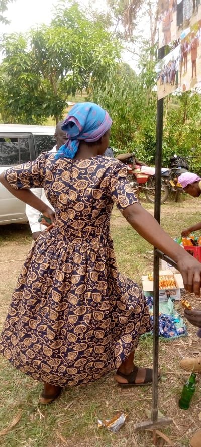 A woman in a blue head scarf and a long, black-and-gold floral dress stands in a grassy area next to a metal pole and a white car. She is facing the camera, but looking to the right, and is holding her right hand on the pole, while her left arm hangs down by her side. A man in a white shirt stands behind her, looking to the right. In the distance, a motorcycle is parked, and a woman in a pink headscarf can be seen sitting on the ground. Several items, including a large bottle of soda, are scattered on the ground in front of the woman. This scene takes place in Burundu, Kenya, at the coordinates provided.