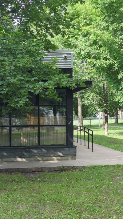 A modern building with a corrugated metal exterior and large glass windows stands beside a paved pathway. Lush green trees and grass surround the structure, suggesting an outdoor setting in a park or residential area. The time of day appears to be daytime, with bright sunlight casting shadows. The weather seems to be pleasant, as indicated by the vibrant greenery and clear visibility. The scene is calm and serene, with no people visible, and the overall atmosphere is one of peacefulness and natural beauty. The location is identified as Montreal, Canada, which hints at a temperate climate and perhaps an urban or suburban environment with ample green spaces.