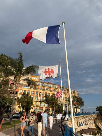 The image depicts a cloudy day in Nice, France, with a prominent French flag waving in the foreground. Behind the flag, a building with yellow facades and balconies, identified by signage as "HOTEL SUISSE", stands tall. To the left of the hotel, a lush green palm tree arches over the scene, and further left, a hillside is covered in vegetation.
Several people are visible on a paved walkway. On the right side of the image, a wall displays the word "CASTEL" in large blue letters, suggesting this is the Castel Plage area. Another flag, white with a red eagle emblem, is also flying, likely representing the city of Nice. An American flag is also visible flying on a flagpole.
The people on the walkway are casually dressed, with some walking and others standing. One individual with a cast on their leg is seated on a blue railing. The overall atmosphere suggests a tourist area with a mix of locals and visitors enjoying the seaside promenade. The lighting suggests it is late afternoon.