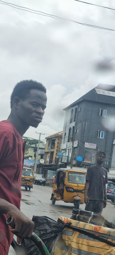 The image depicts a street scene in Alagbede, Nigeria, with a cloudy sky suggesting an overcast day. The foreground is dominated by the shoulder and arm of a person wearing a maroon shirt, holding onto a metal bar. Their gaze is directed upwards and to the right.

In the mid-ground, a yellow auto-rickshaw is parked. Behind it, a man in a dark grey t-shirt stands looking towards the camera. Further in the background, several buildings line the street. One building is a multi-story structure with a dark facade and blue-framed windows. Another building appears to be older, with a lighter, yellowish exterior.

Several signs are visible on the buildings. One sign reads "KADOS". Other signs are less legible due to the image quality and distance. There are also utility poles with wires crisscrossing the scene. The presence of auto-rickshaws, the architecture of the buildings, and the general streetscape are characteristic of many urban areas in Nigeria. The overall impression is of a bustling, everyday street scene in a Nigerian town or city.