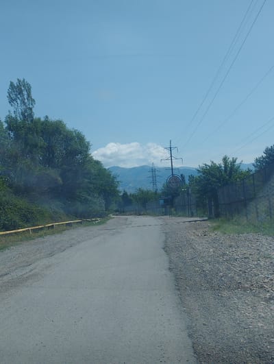 The image depicts a paved road in Namdanak, Uzbekistan, on a clear day with a blue sky and scattered clouds. The road is flanked by a gravel shoulder on the right and a grassy area with a yellow guardrail on the left. In the background, there are lush green trees, mountains, and power lines with electricity towers. A circular sign with the text "RANO ECO-VILLAGE" and a "P" parking symbol is visible on the right side of the road. The scene appears to be an entrance to a rural or eco-tourism area.