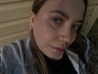 The media file is a close-up, low-angle portrait of a young woman with light skin, brown hair, and blue-green eyes. She is wearing a light blue denim jacket over a black garment and has several piercings, including a hoop in her nose and a larger hoop in her left ear, along with a smaller stud. Her lips are glossed a pale pink, and she has subtle makeup on. The image appears to have been taken outdoors during daylight, with natural lighting. The background consists of a textured wooden surface, possibly siding on a building, and a patch of dry grass and concrete. The woman's gaze is directed towards the camera, and her expression is neutral. The overall impression is casual and personal.