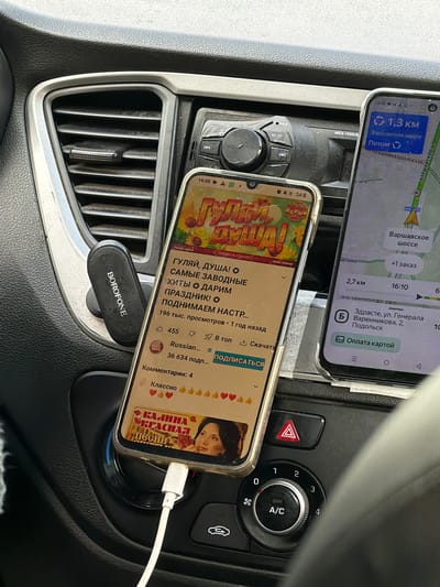 The image shows the dashboard of a car. Two smartphones are mounted in a car vent holder, one above the other. The top phone displays a navigation app with directions to "Варенникова, 2, Подольск", indicating an address on ulitsa Generala in Podolsk, Russia. The navigation app also shows a distance of 1.3km to Varshavskoye shosse. The bottom phone is playing a YouTube video with a title in Russian that translates to "Walk, Soul! The Most Catchy Hits We Give You a Holiday!". A "BOROFONE" branded phone holder is attached to the car vent. The lower phone is connected to a white charging cable. The car's hazard lights button, A/C control knob, and a couple of unidentified buttons are visible on the right side of the dashboard. The vent is surrounded by plastic, which is covered in dust.