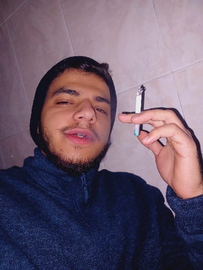 A man lies on a tiled surface wearing a black beanie and a dark blue sweater. He holds a lit cigarette in his right hand, positioned near his head. He has a beard and mustache and is looking toward the camera with slightly narrowed eyes and an open mouth. The room is illuminated with a soft light.