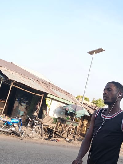 The image depicts an outdoor scene in Gwagwarwa, Nigeria, under a bright, clear sky. In the foreground, a dark-skinned male with a short beard and short hair is visible from the chest up, positioned on the right side of the frame. He wears a black tank top with white and red trim around the collar and armholes, and has white wired earbuds in both ears. His mouth is slightly open, and he appears to be looking towards the left.
The background features a street or unpaved road in the lower left, where a blue motorcycle and a white bicycle are parked. Directly behind them and extending across the mid-ground are several single-story structures with weathered, corrugated metal roofs. These buildings appear to be shops or stalls. One structure to the left has an open front with a dark interior, and a person is partially visible inside. A green sign with "OG" text is visible on the wall of one building. To the right of this, a makeshift stall constructed from wooden planks displays multiple small, dark-colored bottles, possibly containing liquids, under a partially collapsed green and black umbrella.
Dominating the upper-right background is a tall, slender metal pole topped with a rectangular solar panel and integrated light fixture, indicative of a solar-powered street light. The overall impression is a bustling, informal commercial street.