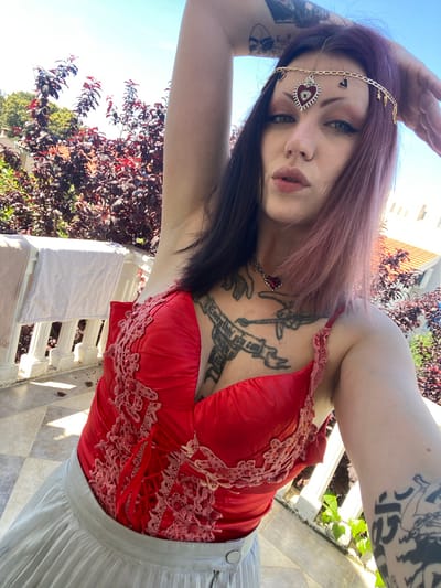 A young woman with long, dark roots blending into purple-pink hair poses for a selfie in bright daylight. Her gaze is direct, accentuated by prominent dark eyeliner and light-colored eyes, and she has a philtrum piercing. She wears a golden headpiece adorned with small charms like a heart, stars, and a crescent moon, culminating in a central red heart-shaped evil eye pendant on her forehead. A matching red heart pendant hangs from her neck.
She is dressed in a vibrant red top, possibly a corset, featuring intricate light pink lace and beadwork on the front and visible lacing at the back. A light grey pleated skirt completes her outfit. Her left arm is covered in a dark, intricate tattoo sleeve depicting figures and patterns, while her chest features additional tattoos, including a banner that partially reads "EVERYTHING YOU CAN". Her right arm is raised, partially obscuring the top of her head as she takes the photo.
The scene is an outdoor setting, likely a balcony or terrace in Kunje, Montenegro, under a clear blue sky. Lush foliage with dark reddish-purple and green leaves forms a natural backdrop. A white balustrade or railing and a light-colored tiled floor are visible to her right. In the upper background, a light-colored building with an orange-tiled roof can be seen. The bright sunlight indicates it is daytime, casting clear shadows.