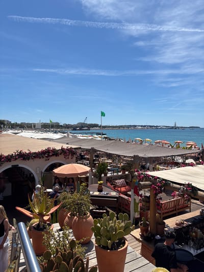 This image shows a coastal scene in Cannes, France. The right side of the frame is a restaurant or beach club with potted cacti, tables, and seating. People are present, some seated and some moving. The middle ground shows a beach with parasols and people near the water's edge. Beyond the beach, the blue sea stretches to the horizon, where a distant coastline is visible. The left side of the image shows a clear blue sky with contrails. A green flag is flying near the beach. The perspective is taken from an elevated position, looking down towards the beach and sea.