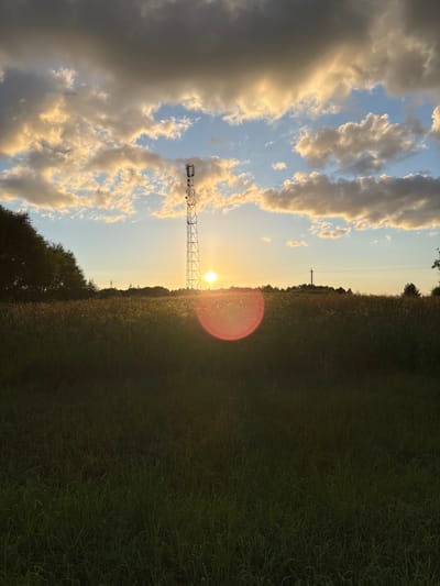 The image depicts a sunset over a rural landscape in Відзаўскі сельскі Савет, Belarus. A tall cellular tower stands prominently in the center-right of the frame, silhouetted against the setting sun. The sky is a gradient of blue and orange, with dramatic clouds tinged yellow by the sunlight. A lens flare, appearing as a reddish-pink circle, is visible in the lower-center of the image, originating from the sun. In the foreground, a field of tall, dark green grass extends across the bottom half of the image. To the left, a dense line of trees forms a dark silhouette against the sky. The overall scene is one of natural beauty and a touch of man-made infrastructure against a backdrop of the day's end.