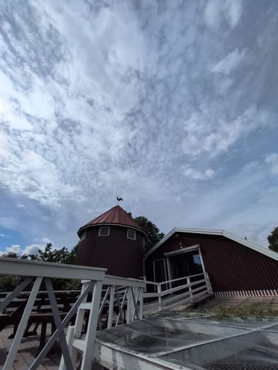 The image shows a view of two rustic, red wooden buildings against a sky filled with scattered white clouds. The building on the left is a cylindrical structure with a conical roof, topped with a black weather vane in the shape of a rooster. It has two small, louvered windows on the visible side. The building on the right has an arched, sloping roof and a large glass door. A white wooden railing and a pathway with a metal grate are in the foreground, with picnic tables visible behind the railing. Trees and foliage surround the buildings. The overall impression is one of a rural or park-like setting. This image was taken at the Seurasaari Open-Air Museum in Helsinki, Finland.