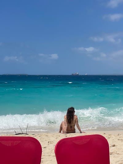 The scene depicts a beach in Zanzibar, Tanzania, under a clear blue sky with scattered white clouds. In the immediate foreground, the backs of two red beach chairs are partially visible, framing the view. A single individual, identified as female, is seated on the light-colored sand, facing away from the camera and looking out towards the ocean. She is wearing a dark bikini top, and her long, dark hair is visible. A white wave is breaking directly in front of her, and small pieces of driftwood or debris are present on the sand to her left. The ocean extends beyond the beach, exhibiting a clear gradient from turquoise near the shore to a darker blue further out. On the distant horizon, a large cargo vessel is visible, along with a small white sailboat to its right. A low, distant landmass or island is discernible on the far left horizon. No direct interactions between individuals are depicted.
