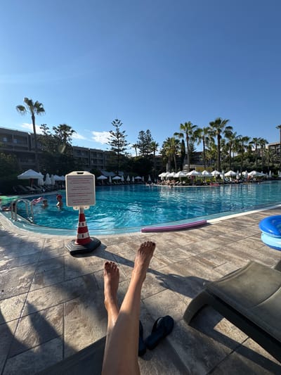 The image shows a resort swimming pool on a sunny day in Manavgat, Turkey. In the foreground, a person's legs and feet are visible, stretched out on a lounge chair. The feet are bare, with dark red nail polish. A pair of black flip-flops are placed on the tiled ground next to the chair.
The swimming pool is large and blue, with clear water. Several people are in the pool, some swimming and others relaxing. There are lounge chairs with white umbrellas surrounding the pool, and a building complex with balconies is visible in the background. Palm trees and other greenery are scattered throughout the scene.
A red and white striped cone with a sign attached to it stands at the edge of the pool. The sign contains text in multiple languages, including Turkish and English, warning that there is "NO LIFEGUARD ON DUTY" and "SWIMMING IS AT YOUR OWN RISK." A pink pool noodle and a blue inflatable ring are floating in the pool. The ground around the pool is paved with large, square tiles, and is wet in some areas, reflecting the sunlight. The sky is clear and blue.