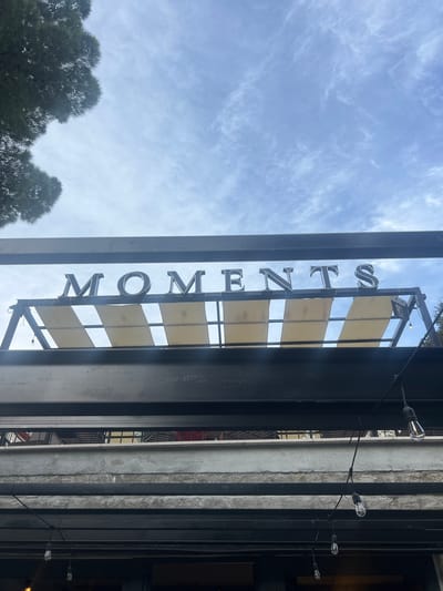 The image shows a close-up view of a sign spelling out "MOMENTS" in large, dark, three-dimensional letters, mounted on a metal frame above an awning. The awning is made of tan fabric panels stretched across a metal structure. The sign is silhouetted against a sky with scattered white clouds. In the upper left corner, a portion of a dark green tree is visible. Below the awning and sign, multiple layers of dark metal beams are visible, suggesting the structure of a building or patio cover. A string of exposed filament light bulbs hangs from one of the metal beams, with several bulbs illuminated and others not. The lighting suggests it may be late afternoon or early evening. The scene appears to be an outdoor setting, possibly a restaurant or cafe, in Dobrota, Montenegro.