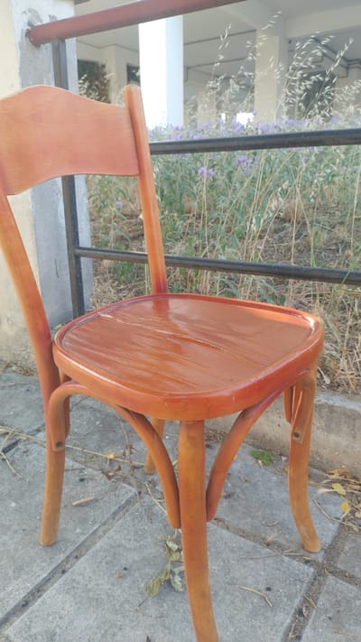 An outdoor medium shot captures a close-up of a worn, light brown wooden chair. The chair's seat is made of bent wood and shows scuff marks and scratches. The chair has four curved legs, and its backrest is visible in the upper left quadrant of the image. The chair is positioned on a paved surface, possibly a sidewalk, composed of large, rectangular grey tiles with visible cracks and debris. In the background, a dark metal railing separates the foreground from an area filled with dry grass and small purple flowers. Behind the railing, there is a blurred view of a building with white pillars and a red-colored railing overhead. The setting appears to be in the city of Perea, Greece. There are no people present in the image, and no actions are being depicted.