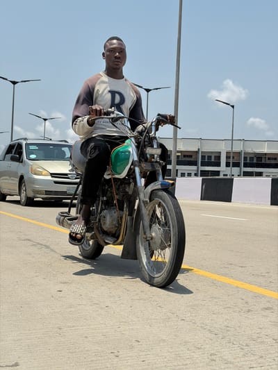 The image displays a young adult male seated on a motorcycle on a paved road in Alaguntan, Nigeria. The male, with dark skin and a short haircut, wears a grey and dark-sleeved raglan shirt featuring a prominent white "R" emblem, black trousers, and black-and-white striped slide sandals. He grips the motorcycle's handlebars and looks directly forward.
The motorcycle is an older model, distinguished by a green and white fuel tank, exposed engine components, spoked wheels, and a visible front fender. A silver Toyota Sienna minivan, bearing the license plate "KRD 659 FK", is positioned behind the motorcycle to the left. The road surface is concrete or asphalt, marked with a solid yellow lane line. A black and white striped concrete barrier runs along the right side of the road. In the background, modern streetlights with flat fixtures are visible on tall poles, alongside multi-story buildings. The sky is clear blue with minimal clouds, indicating bright daylight that casts distinct shadows.