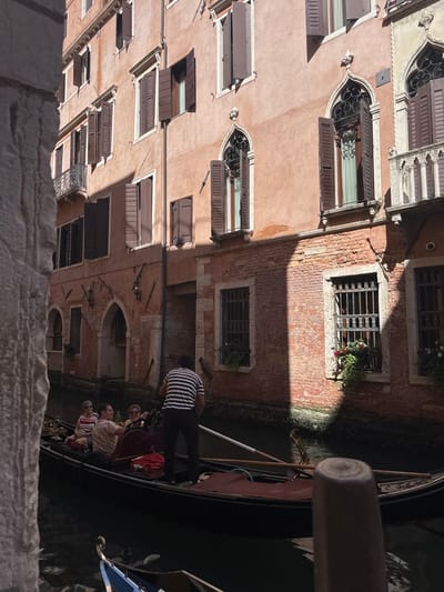The image captures a scene in Venice, Italy, featuring a gondola carrying several people as it navigates a narrow canal. The gondola is dark green with ornate gold trim. A gondolier, wearing a black and white striped shirt and black pants, stands at the back, steering with a long oar. Inside the gondola, five passengers are seated, their faces visible. Sunlight casts strong shadows on the buildings lining the canal, suggesting it is either morning or late afternoon.
The buildings are made of brick and plaster, with a warm, faded orange hue. They feature numerous windows, many with closed wooden shutters. Some windows have decorative iron grills, and a few have small potted plants with red and white flowers. Architectural details like arched doorways and intricate stonework are visible. The canal water is dark and reflects the shadows. The overall atmosphere is tranquil and evokes the classic Venetian experience. The image is shot from a slightly elevated perspective, possibly from a bridge or balcony, looking down onto the canal and gondola.