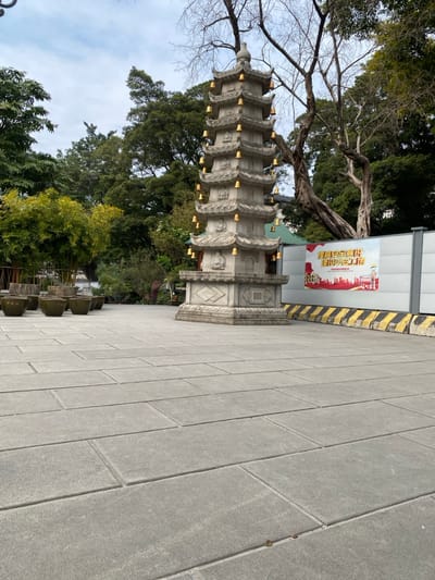 This image captures an outdoor scene featuring a multi-tiered stone pagoda, lush greenery, and a paved walkway. The pagoda, made of light grey stone, stands centrally and features intricate carvings and small golden bells or ornaments hanging from the eaves of each tier. Its base is broad and also carved with decorative patterns.

Surrounding the pagoda are various trees; some have bare branches, suggesting a cooler season, while others are full of vibrant green leaves. In the background, to the right of the pagoda, there is a temporary white wall or barrier with a yellow and black striped base. A large red banner is affixed to this wall. The banner displays Chinese characters, prominently "喜迎二十大" (Xǐ yíng èrshí dà), which translates to "Happily Welcome the 20th National Congress," and "奋进新征程" (Fènjìn xīn zhēngchéng), meaning "Strive forward on a new journey." A stylized red cityscape silhouette is also visible on the banner, along with a blurred series of zeros that appears to be a phone number.

The foreground and right side of the image show a wide paved area composed of rectangular grey tiles. A line of dark, square planters is arranged along the edge of this paved area near the base of the pagoda. The sky overhead is overcast, indicating a cloudy daytime setting. The scene evokes a tranquil, possibly historical, public space or temple ground in Guangzhou City, China.