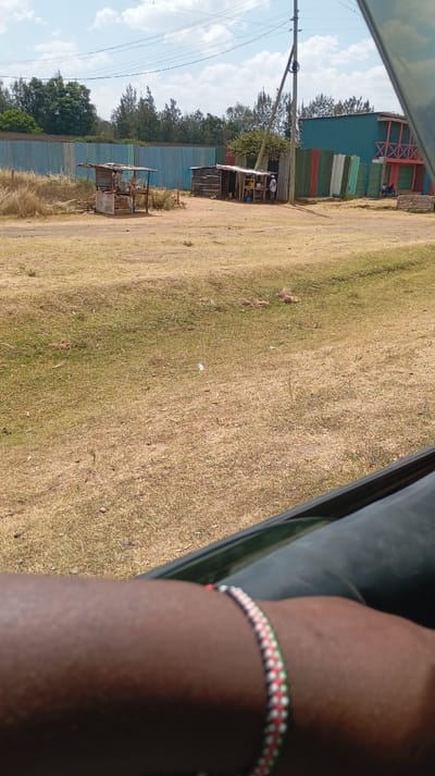 A sunny, dusty scene unfolds from what appears to be a vehicle. Dilapidated wooden shacks line a dirt road near fences; the one on the right is multi-colored, the other is blue. Beyond the fences, trees dot the horizon under a bright sky with sparse clouds. In the foreground, part of a person's arm is visible, adorned with a thin, colorful bracelet of white, red, green, and black beads.