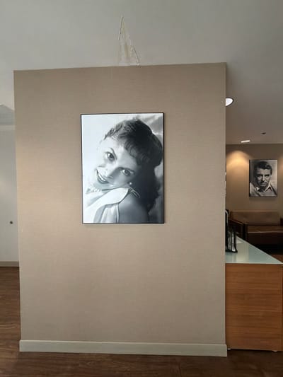 The image depicts an interior scene, likely a hallway or office space, with a neutral-toned wall as the primary background. Dominating the foreground is a framed black and white portrait of Audrey Hepburn, positioned centrally on the wall. To the right, further down the hallway, another framed black and white portrait of Cary Grant is visible. The scene also shows part of a reception desk with a wooden cabinet and a light-colored countertop. The floor is covered with dark wood flooring. In the upper section of the image, a damaged area of the ceiling is apparent. The location is identified as Burbank, United States of America.
