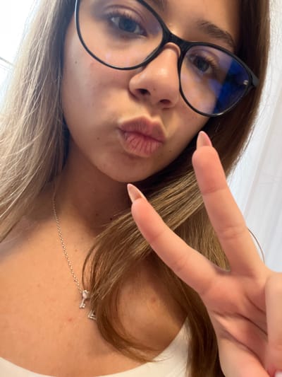 A young woman is depicted in a close-up, self-portrait-style image, taken in Karavelovo, Bulgaria. She has light brown hair, wears black-rimmed glasses, and puckers her lips in a kissing gesture. Her left hand is raised, with the index and middle fingers extended to form a peace sign, revealing long, light-pink almond-shaped fingernails. She wears a silver chain necklace adorned with two small charms: one resembling a key and the other an initial, possibly 'A'. Her skin exhibits some minor textural variations. The background is softly blurred, showing indistinct light-colored elements, indicative of an indoor setting.