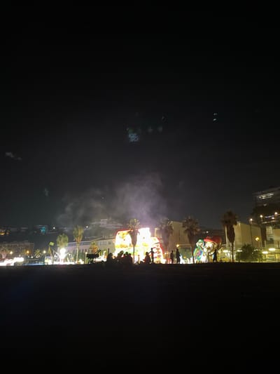 The image depicts a nighttime scene, likely a fair or festival, in Canet de Mar, Spain. The orientation of the image is rotated, with the buildings and activity appearing on the right side and a large dark area on the left.

The subjects are a crowd of silhouetted people mingling amongst various attractions. Prominently featured are inflatable structures, one resembling a large character with a red hat and another a colorful bounce house. Illuminated stalls and rides create a vibrant, festive atmosphere with bright lights. Palm trees are visible, suggesting a warm climate. Buildings line the background, illuminated by the event's lights and streetlights.

The activity appears to be a nighttime gathering at an amusement event. People are walking around, observing the attractions, and likely participating in the festivities. Wisps of smoke or mist are present, adding to the atmospheric lighting.

Notable details include the time of day, which is clearly night due to the darkness and artificial lighting. The weather appears calm. Location cues, like palm trees and architecture, along with the provided context, point to Canet de Mar. The mood conveyed is one of lively celebration and enjoyment.

There is no visible text in the image.
