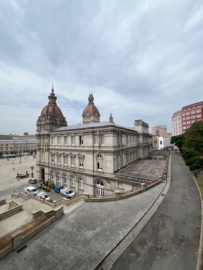 This is a vertical, eye-level, medium shot of a street in A Coruña, Spain, with a focus on an ornate, grey building featuring classical architectural elements like columns, arches, and domes. The building has numerous windows and decorative detailing along its facade. In front of the building, a paved walkway with cobblestone detailing transitions to a paved road. Several white vans and a blue truck are parked along the street, along with a small group of people, some wearing yellow safety vests, suggesting some sort of work or maintenance is occurring. Further down the street, other buildings are visible, including a taller, reddish-brown building. The sky is overcast with visible clouds, indicating it is likely daytime but not particularly sunny. The scene appears to be a public space, possibly a plaza or a street in a city center. The overall impression is one of an urban environment with historical architecture.