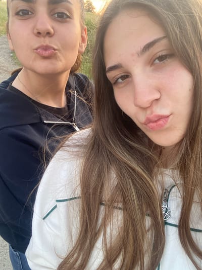 The image captures two young adult females posing outdoors in Debelt, Bulgaria, under illumination consistent with late afternoon or early evening. The female positioned in the left background has brown hair, brown eyes, and a light complexion with minor facial blemishes, her lips pursed. She wears a dark-colored zippered hoodie over a darker undergarment, featuring a visible silver chain necklace. The female in the right foreground has long, brown hair with lighter highlights, a light complexion with minor blemishes and freckles, and her lips are also pursed. She is dressed in an off-white, long-sleeved top with thin dark-green stripe details and a silver zipper pull displaying a circular, stylized emblem. The background is out of focus, revealing an unpaved ground surface with patches of dry grass and distant trees under a warm, orange-tinted sky. Both individuals are engaged in a shared pose for the photograph.