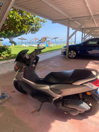 A grey Yamaha NMAX scooter is parked under a covered structure, casting a shadow on the red ground. Behind the scooter, a dark blue sedan is parked. In the background, there is a well-maintained lawn with several sun loungers and straw umbrellas. Beyond the lawn, the azure sea and a clear blue sky are visible. A lush green tree with large leaves stands to the left, and a stone path with a blue hose reel is in the foreground. The location appears to be a coastal resort area in Estepona, Spain, likely a hotel or residential complex given the amenities.
