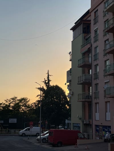 The image captures a street scene in Nesebar, Bulgaria, during what appears to be sunset. The primary subject is a multi-story building with a light pink facade, featuring numerous balconies with metal railings and multiple windows. Adjacent to it is another section of the building painted in a pale green hue. Several satellite dishes are mounted on the building.

In the foreground, a street is visible with parked vehicles. A prominent red van is parked on the right side of the street. Behind it, a white van and a white car are also visible. A red stop sign is placed on a pole near the vehicles. There are also metal barriers on the sidewalk.

Trees with dark green foliage line the street, partially obscuring the buildings and the sky. The sky itself is a soft gradient of pale blue and warm yellow, indicative of sunset. A thin, dark line, possibly a wire or a clothesline, stretches across the sky from the upper left towards the center. A single bird is visible in flight against the sky.

One sign on the building has text that appears to be in Cyrillic, but it is difficult to read clearly. Another sign further down the street also appears to have Cyrillic text. The overall mood is peaceful and tranquil, with the warm light of sunset casting a gentle glow on the scene.