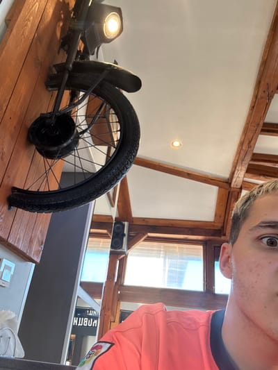 A close-up, slightly tilted shot captures a young man's right eye and upper face, as well as a motorcycle wheel mounted on a wooden wall. The man, with short, light-colored hair and brown eyes, is wearing a bright pink shirt. The motorcycle wheel, black with spokes, is positioned vertically on a wooden planked wall. A lamp attached to the motorcycle's handlebars, above the wheel, emits a bright yellow light. The background shows exposed wooden beams and a white ceiling, with a window visible further back. The scene appears to be indoors, possibly a cafe or restaurant, given the decor.