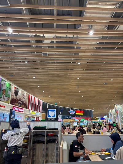 This image captures a vibrant and spacious food court, likely within a mall, in Bekasi Regency, Indonesia. The scene is dominated by a high ceiling featuring numerous light-colored wooden slats arranged in parallel, creating a modern, airy feel. Exposed ventilation ducts and recessed lighting fixtures are visible between the slats. The floor below also appears to be made of similar light-colored wood.

Numerous people are present, engaged in dining and socializing. In the foreground, a man wearing a black shirt with an Indonesian flag emblem is seated at a table, looking down at his food. Another person is partially visible across from him. Further into the food court, many more patrons are seated at various tables and food stalls. On the right side of the frame, a person wearing a plaid jacket with "FOOD COURT" emblazoned on the back is seen near a multi-tiered cart, possibly for collecting used trays.

The food court features several food vendors with brightly lit signs. A prominent black pillar-like structure in the mid-ground displays a white and blue "RETURN" sign, featuring an icon of a bowl and spoon, indicating a drop-off point for trays.

Visible text on various signs includes: "MULTI", "AKIHABARA", "RETURN", "Golden Onion", "Chicken Shabu", "from UAE to the WORLD", and "TLC Chicken". The lighting suggests it is daytime or a well-lit interior. The overall atmosphere appears busy and casual, characteristic of a public dining area.