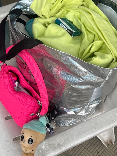 A close-up, eye-level shot shows the contents of an open, silver-lined tote bag. The bag contains crumpled neon-yellow fabric, possibly clothing, and a small teal box of "TEREA" brand tobacco sticks. A bright pink zippered pouch hangs from a black strap connected to the tote bag. Inside the pouch, a pink object, possibly a lip balm or pen, is visible. A small, plush toy with a light brown body, a light blue fuzzy hat, and large black eyes is attached to a keychain hanging from the pink pouch. The tote bag and pouch rest on a white surface, which appears to be part of an outdoor seating area, possibly a chair or bench, with a patterned stone or tile floor visible in the background. The scene is well-lit, suggesting it is daytime. The context suggests a casual outing or preparation for travel, with the items likely belonging to one individual. There is no indication of the specific location within Kranevo, Bulgaria, other than the general context of an outdoor setting.