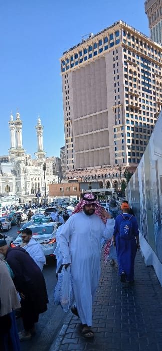 The image displays a bustling urban street scene in Makkah Al Mukarramah, Saudi Arabia, under a clear blue sky. In the foreground, a male subject wearing a white thobe, a red and white checkered ghutra secured by an agal, and sunglasses walks towards the viewer on a dark-tiled sidewalk, carrying a white plastic bag in his left hand. To his right, a second individual, dressed in a blue abaya with an orange head covering and a blue backpack, walks away from the viewer. The right side of the sidewalk is bordered by a light-colored construction hoarding. The mid-ground reveals a multi-lane roadway heavily congested with vehicular traffic, predominantly white and light-colored sedans. In the background, to the left, two distinct, ornate white minarets with multiple balconies, characteristic of the Grand Mosque, are prominently visible. A large, multi-story, light-colored modern building with numerous blue-tinted windows dominates the right background, with additional urban structures extending into the distance. Numerous other pedestrians are visible on the sidewalks and within the street area.