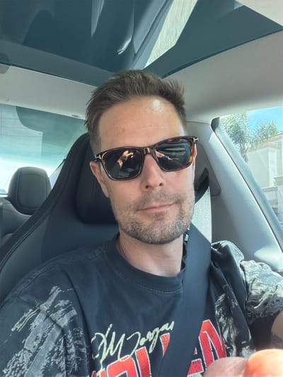 A Caucasian male, appearing to be in his mid-30s to early 40s, with short brown hair and light stubble, is depicted in a selfie taken from the driver's seat of a vehicle. He is wearing dark tortoiseshell sunglasses, which reflect his right hand holding a smartphone, indicating he is taking the photograph. He is wearing a dark t-shirt with a light-colored graphic design on the shoulders and a large red and yellow graphic on the chest, partially obscured by a black seatbelt fastened across his body.
The car's interior features black leather seats, a light gray fabric headliner, and a panoramic glass roof, which reflects elements of the sky and light. The headrest of the passenger seat is visible behind him.
Through the vehicle's windows and in reflections, a clear blue sky, bright sunlight, and several tall palm trees are prominent. On the right side, a light-colored, multi-story building with architectural features, possibly stucco or stone, is partially visible. The overall environment, characterized by palm trees, specific architectural styles, and clear sunny weather, is highly consistent with many areas within the city of Los Angeles, California, United States of America.