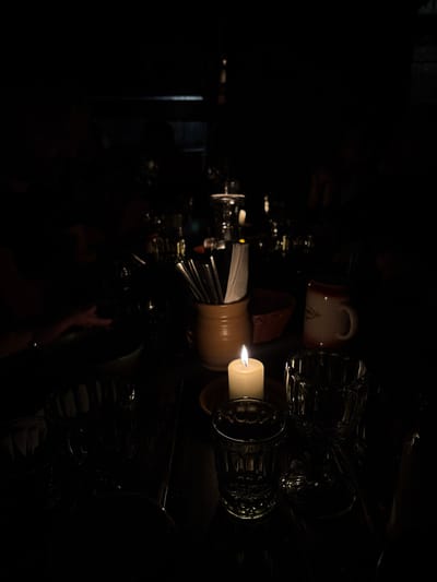This image captures a dimly lit indoor dining scene within a restaurant or pub in the Capital City of Prague, Czechia. The central focus is a lit, cream-colored pillar candle resting on a small, dark terracotta-colored saucer on a dark wooden table. To the candle's immediate left, a light brown stoneware crock holds an assortment of silver-colored cutlery and folded white paper napkins. Partially visible behind these items is a woven basket. To the candle's right, a light-colored ceramic mug with a handle and a subtle decorative pattern is present. In the foreground, two clear, textured glass tumblers are positioned on the table surface. The background is predominantly dark and out of focus, revealing blurred shapes indicative of other tables and patrons. On the left side of the table, a person's hand and forearm are faintly visible next to a light-colored bowl, suggesting a diner seated at the table. The primary illumination originates solely from the candle flame, contributing to a dark and intimate ambiance.